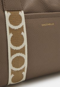 Bruin leer met textuur tas met een beige strap met een cirkelpatroon en een gouden reliëf logo met de tekst "COCCINELLE."
