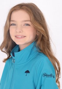 Giacca softshell teal con colletto alto, caratterizzata da un logo ombrello navy e testo ricamato. Texture liscia e design aderente.
