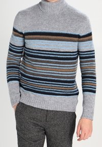 Šedý proužkovaný svetr s vysokým límcem, zdobený horizontálními liniemi v tmavě hnědé, modré a námořnické modři. Měkká pletená textura s žebrovanými manžetami.