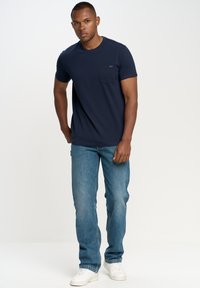 Granatowa koszulka z kieszonką na piersi, w połączeniu z jasnoniebieskimi dżinsami. Model w białych trampkach, stojący na jednolitym tle.