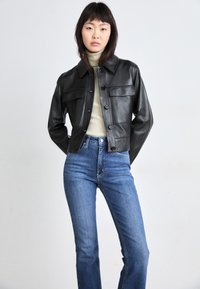 Giacca in pelle nera corta con bottoni a pressione, due tasche sul petto e colletto, abbinata a jeans blu classici a vita alta.