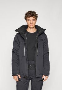 Giacca nera isolante con cappuccio, dotata di tasche con zip, tessuto texturizzato e vestibilità rilassata. Indossata con una maglietta nera e pantaloni.