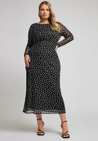 Musta pallokuvioinen mekko, jossa on pitkät läpikuultavat hihat, tiukka yläosa ja liehuva helma. Mekossa on pyöreä pääntie ja nilkkapituinen malli.