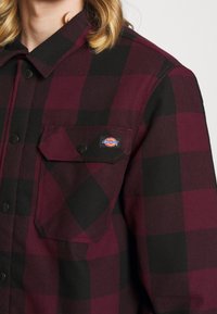 Chemise en flanelle à carreaux rouges et noirs avec un bouton devant, deux poches poitrine et une petite étiquette de marque sur la poche gauche.