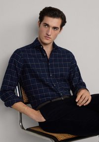 Camicia a quadri blu navy in cotone con colletto button-down. Modello seduto, indossando pantaloni scuri, che mostra una vestibilità rilassata e uno stile informale.