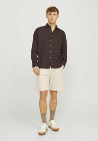 Dunkelbraunes Hemd mit Knopfverschluss und Kragen, beige Shorts, braune Socken und weiße Sneakers mit braunen Sohlen. Das Modell steht vor einem neutralen Hintergrund.