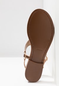 Semelle plate de sandale marron avec talon texturé et marque "ALDO" embossée près du centre, montrant une partie de la bride dorée et de la boucle.