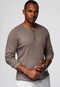 Camisa henley de manga larga con acanalados en tono marrón tierra, con cinco botones en el cuello. Presenta un diseño ajustado y una tela texturizada.