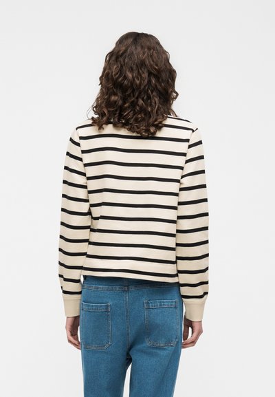 Personne aux cheveux bouclés portant une chemise beige à manches longues avec des rayures horizontales noires et un jean bleu, tournée dos à la caméra.