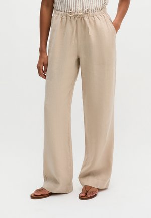 Pantalon beige à larges jambes avec taille élastique et cordon, porté avec des sandales plates marron et une chemise rayée rentrée.