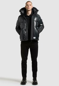 Zwarte gewatteerde jas met een glanzende afwerking, voorzien van een grote capuchon en ritssluiting. Draagt over een zwarte hoodie, in combinatie met zwarte broeken en laarzen.