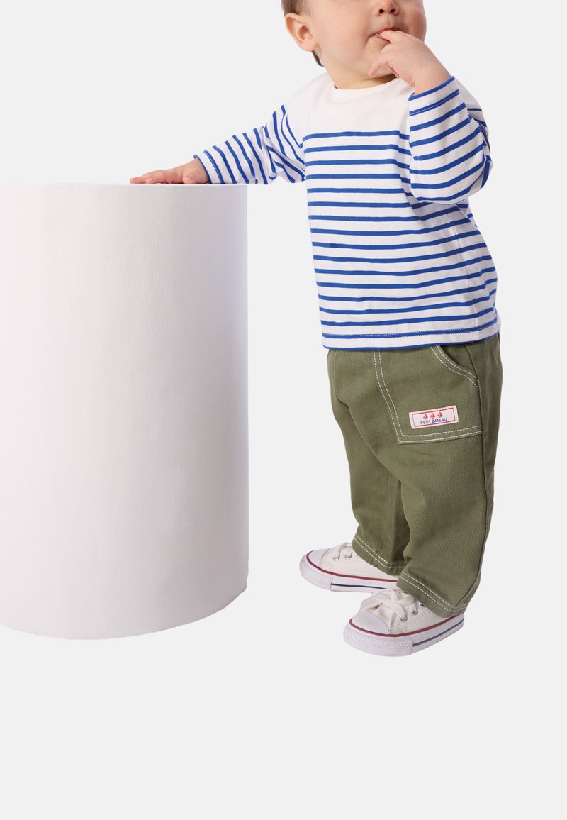 Ριγέ πουκάμισο με μακριά μανίκια σε λευκό και μπλε, παντελόνι ελιάς και λευκά αθλητικά παπούτσια με κόκκινες λεπτομέρειες. Το χέρι ενός παιδιού ακουμπά επιφάνεια κυλινδρικού σχήματος.