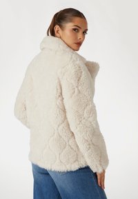 Kreminės spalvos dirbtinio kailio striukė su puriu paviršiumi, turinti deimanto raštą, aukštą apykaklę ir laisvą siluetą. Matomos džinsinės kelnės.