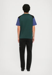 Uomo con capelli ricci che indossa una camicia a maniche corte bloccata nei colori verde, blu e nero e pantaloni neri, in piedi di spalle su uno sfondo bianco.