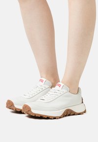 White leather sneakers with lace-up design, rubber outsole, and brown traction pattern; red branding on tongue.