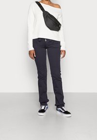 Persoon in een off-shoulder witte trui, zwarte broek, zwarte schoudertas en zwart-wit sneakers die op een grijze vloer staat.