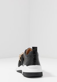 Baskets noires avec une texture crocodile, détail en métal doré et semelle épaisse blanche. Comprend une sangle élastique noire et un tirette à l'arrière.