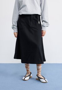 Gonna nera al ginocchio realizzata in un tessuto strutturato, con tasche laterali e vita con coulisse. Abbinata a scarpe stringate bianche e nere.