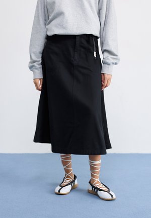 Black, knee-length skirt made of a structured fabric, featuring side pockets and a drawstring waist. Paired with white and black lace-up shoes.
