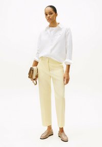 Witte overhemd met knopen, lichtgele op maat gemaakte broek en beige loafers. Houdt een kleine beige handtas vast. Schone lijnen en minimalistisch ontwerp.