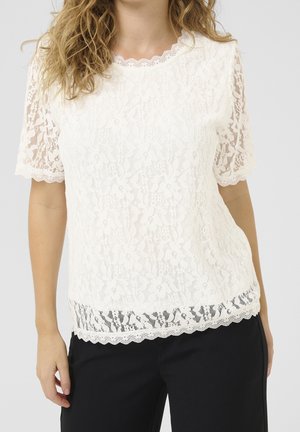 Femme portant un chemisier blanc à manches courtes en dentelle florale avec des bords festonnés et un pantalon noir sur un fond uni.