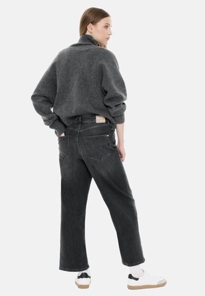 Grauer Pullover mit hohem Kragen, oversized Schnitt, kombiniert mit schwarzen Schlagjeans mit Gesäßtaschen und einem Etikett. Weiße Turnschuhe runden den Look ab.
