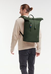 Mochila verde con parte superior enrollable hecha de material impermeable, cuenta con correas ajustables, un bolsillo frontal con cremallera y un asa de transporte.