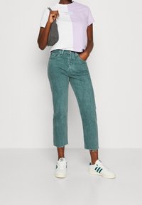 Mujer que lleva una camiseta mitad blanca y mitad lavanda, unos jeans cortos de color teal, zapatillas blancas con rayas verdes y sostiene un bolso de hombro gris.