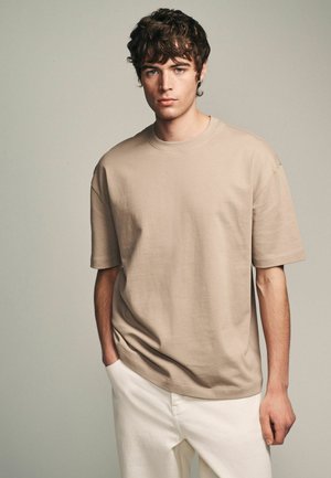 Beige rövid ujjú póló, kerek nyakkal, laza szabású, könnyű pamut anyagból, fehér nadrággal párosítva, minimalista dizájn, minták nélkül.