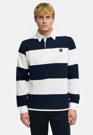 Blonder Mann trägt ein langärmeliges Rugbyhemd mit breiten navyblauen und weißen horizontalen Streifen und einem weißen Kragen, steht vor einem einfarbigen Hintergrund.