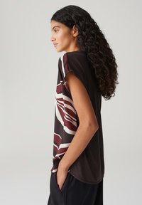 Zwarte korte mouwen top met abstracte, kastanjebruine en witte patronen. Zacht materiaal met een losse pasvorm en afgeronde zoom, zijzak zichtbaar.