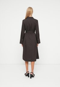 Dunkelbraunes Hemdkleid mit langen Ärmeln, einem Gürtel in der Taille und einem knielangen Saum. Verfügt über umgeschlagene Ärmel und eine glatte Textur. Schwarze Absatzschuhe.