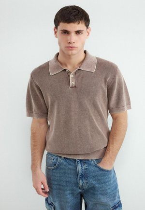 Młody mężczyzna w brązowej, krótkim rękawem, dzianinowej koszulce polo i niebieskich dżinsach, stoi z jedną ręką w kieszeni na jednolitym tle.