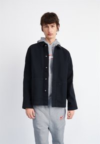 Chaqueta negra de botón con dos bolsillos delanteros, llevada sobre una sudadera gris con un logo rojo, combinada con pantalones de chándal grises.