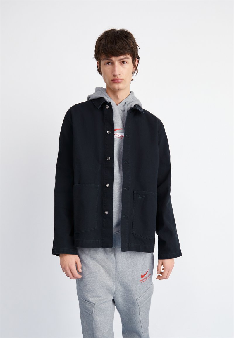 Chaqueta negra de botón con dos bolsillos delanteros, llevada sobre una sudadera gris con un logo rojo, combinada con pantalones de chándal grises.