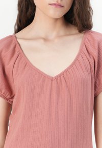 Culoare roz pal, bluză cu decolteu adânc în V, mâneci scurte tip puff și un model texturat cu dungi. Materialul pare moale și ușor.
