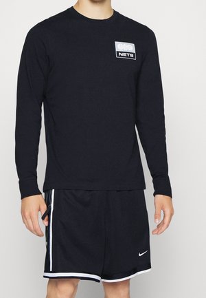 Mand iført sort langærmet skjorte med basketball-logo og sorte Nike sportsshorts med hvide kanter, stående foran en ensfarvet baggrund.