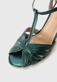 Teal metallic high-heeled sandal with crossed strap detailing and an open toe design, featuring a smooth texture and thin ankle strap.