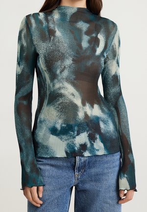 Kvinde iført en langærmet, ribstrikket, halvgennemsigtig top med blå, sort og hvide abstrakte mønstre, kombineret med blå denimjeans.