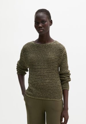 Vrouw met kort haar die een olijfgroene opengebreide trui en bijpassende broek draagt, staand met één hand in haar zak tegen een witte achtergrond.