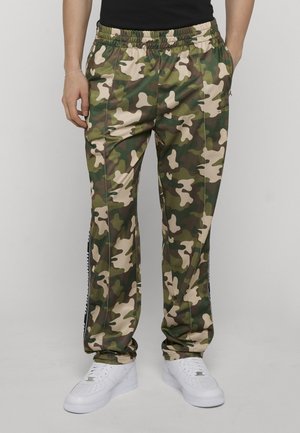 Mand iført grønne og beige camouflagebukser med elastisk talje og hvide sneakers, stående mod en ensfarvet baggrund.