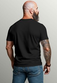 Bald man with a full beard, wearing a black t-shirt and blue jeans, showing his tattooed right arm against a plain light background.