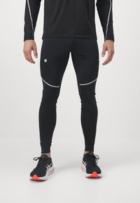 Leggings de sport noirs avec des accents réfléchissants, présentant un tissu lisse et extensible avec un design ajusté. Portés avec des chaussures de course noires.