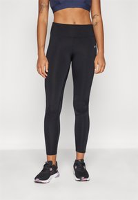 Zwarte legging gemaakt van rekbaar materiaal, met een hoge taille, zijnaden en een subtiele logodetail. Gecombineerd met zwarte sportschoenen.