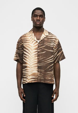 Moški v kratkih rokavih s srajco na gumbe z rjavimi in bež zebrinimi progami, oblečen v črne hlače, na preprostem ozadju.