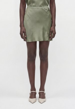 Personne portant une courte jupe en satin vert olive avec une petite fente à l'avant et des escarpins Mary Jane beiges à bouts pointus, debout.