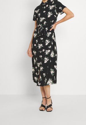 Frau trägt ein schwarzes Midikleid mit pink-weißem Blumenmuster, kurzen Ärmeln, gebundener Taille, Schlitz vorne und schwarzen Sandalen mit Knöchelriemen.