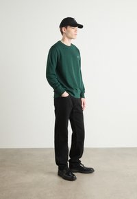 Groene sweatshirt, zwarte broek en zwarte schoenen. De sweatshirt heeft een ronde halslijn en geribbelde manchetten. De pet is zwart met witte tekst.