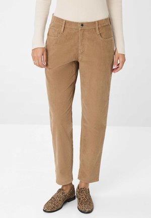 Vrouw in beige corduroy broek en loafers met luipaardprint, staand tegen een effen lichte achtergrond.