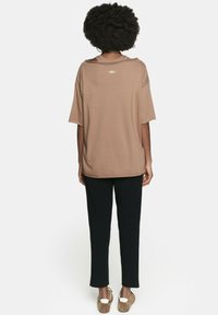 Brązowa bluzka z krótkim rękawem o luźnym kroju, z subtelnym detalem z logo na plecach. Połączona z czarnymi spodniami i jasnobrązowymi butami.